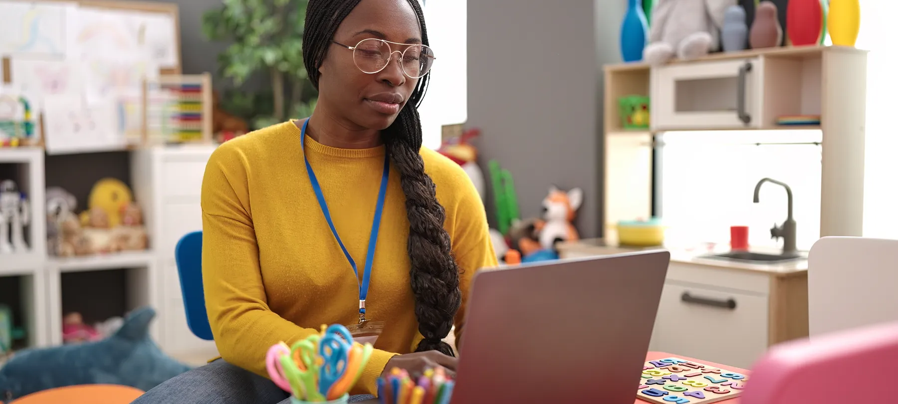 A daycare educator with a lanyard at her laptop in a bright playroom, toys and children’s drawings behind her.