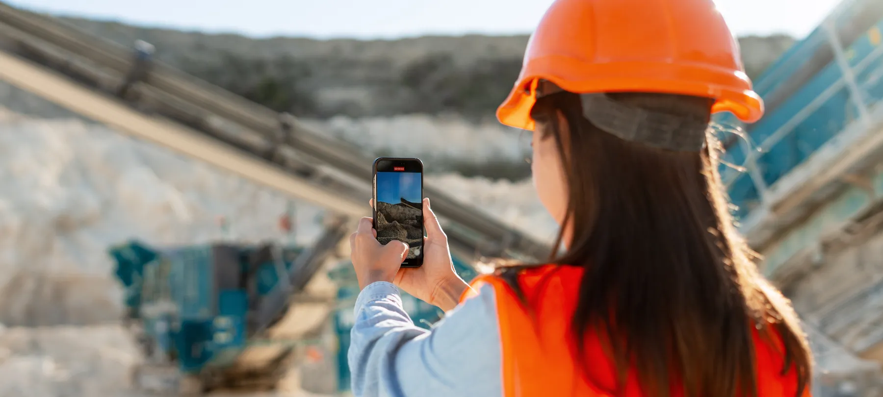 A woman in a hard hat and a hi-vis vest photographing a quarry site with her phone.